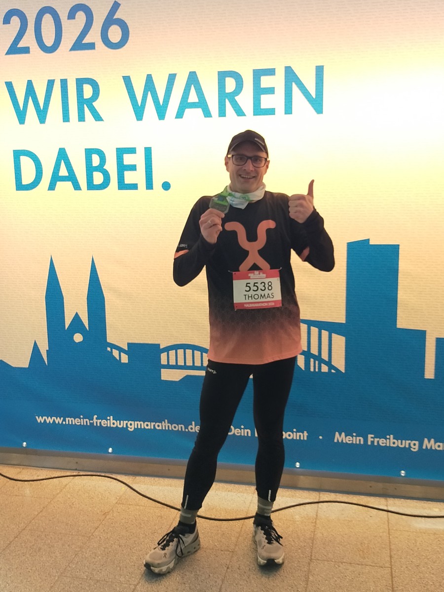 Thomas steht vor der Zielwand mit der Aufschrift Wir waren dabei und hält die Finischer-Medaille in die Kamera.